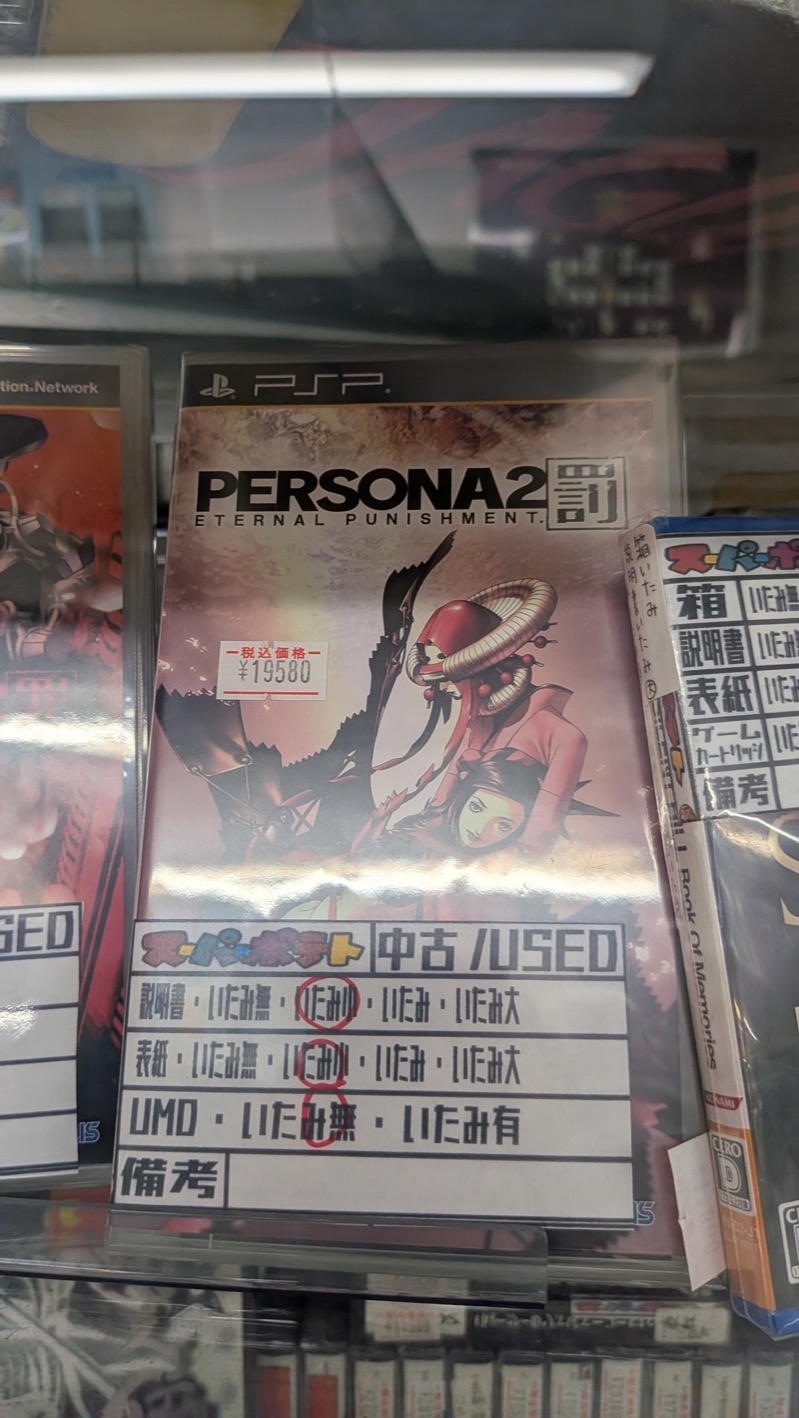 Boxed PSP copy of Persona 2: Eternal Punishment (PERSONA2 罚) in a glass display case at Super Potato, with a &yen;19,580 tax-included price tag and a Super Potato used-condition grading card