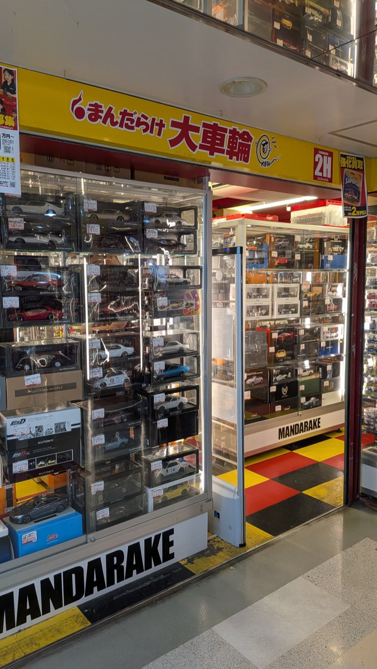 Ladenfront von Mandarake Daisharin, gelbes Schild mit まんだらけ 大車輪 und Wandvitrinen voller verpackter Druckguss-Modellautos