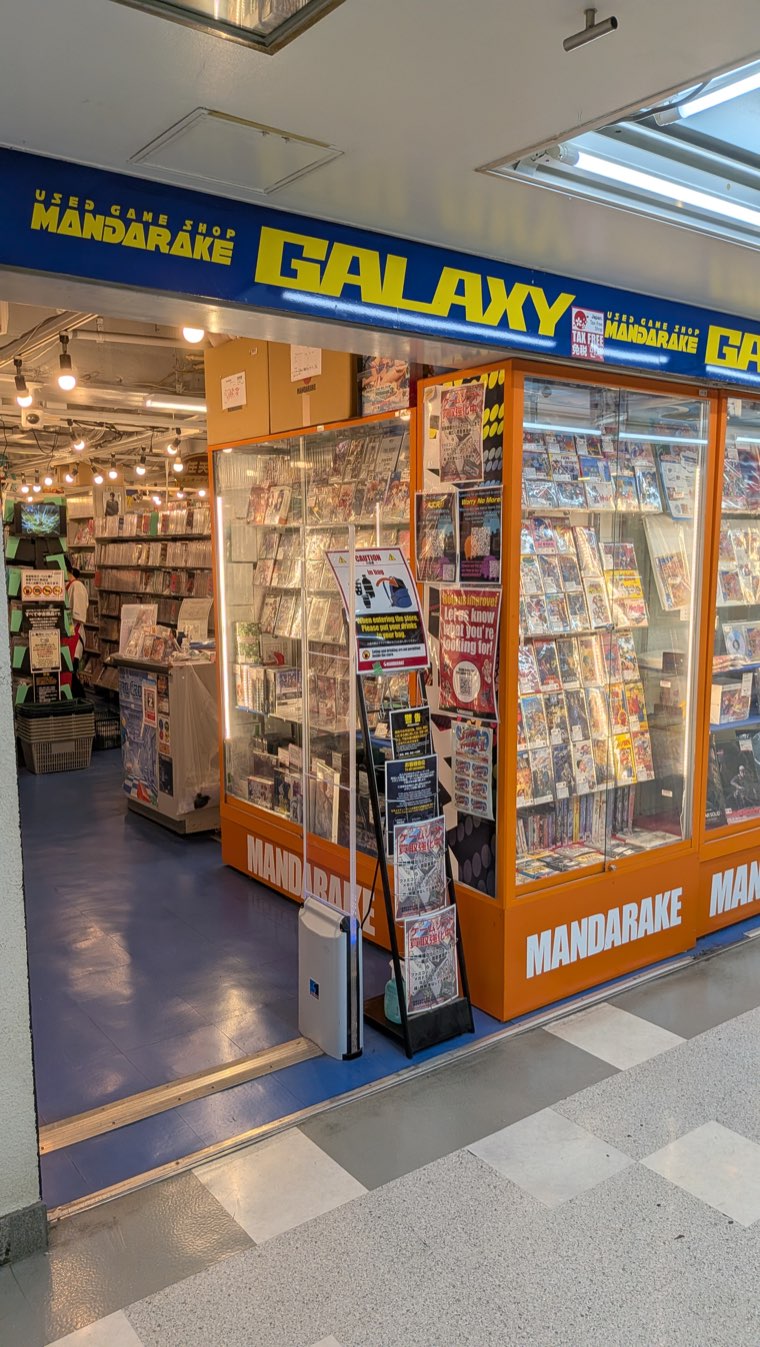 Ladenfront von Mandarake Galaxy in Nakano Broadway, orange-blaues Schild mit USED GAME SHOP MANDARAKE GALAXY und Vitrinen mit verpackten Spielen