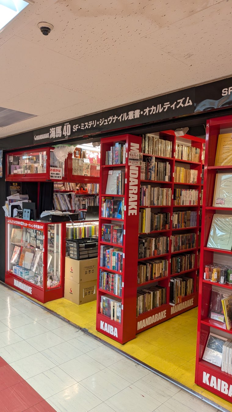 Ladenfront von Mandarake Kaiba im 4. Stock mit einem Schild, das SF-, Krimi-, Jugendbuch- und Okkultismus-Kategorien auf Japanisch auflistet, dahinter hohe Bücherregale