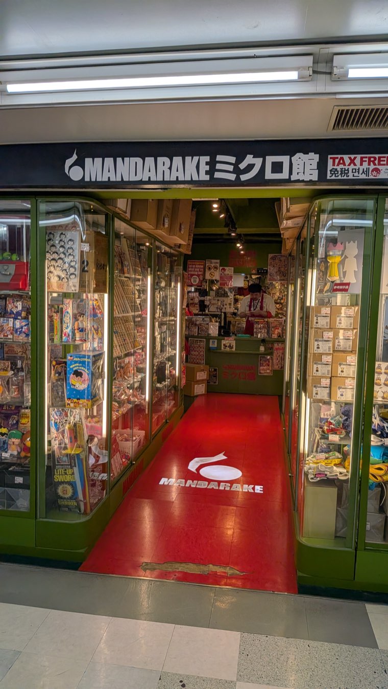 Ladenfront von Mandarake Micro-kan mit grünem Rahmen und rotem Teppich, Vitrinen mit kleinem Vintage-Spielzeug beiderseits des Eingangs