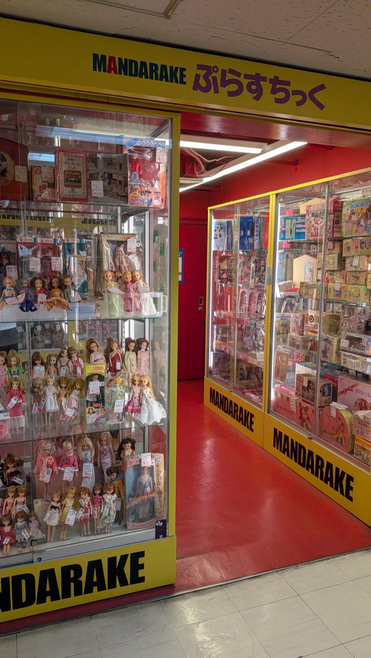 Ladenfront von Mandarake Plastic im 4. Stock mit gelbem Schild ぷらすちっく, Vitrinen mit alten japanischen Modepuppen und verpackten Puppenmöbeln