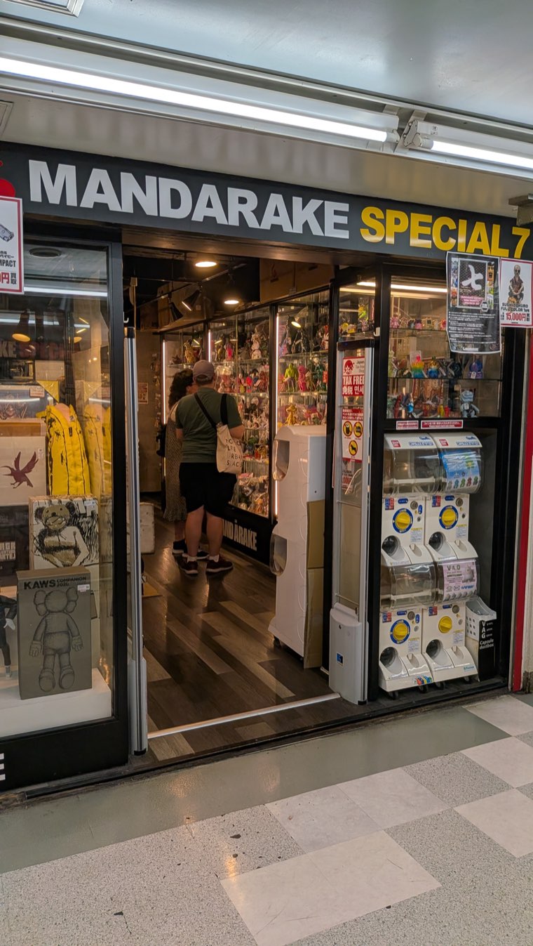 Ladenfront von Mandarake Special 7 mit schwarz-gelbem Schild, Gachapon-Automaten neben einem offenen Eingang und Regalen mit Vinylfiguren im Inneren